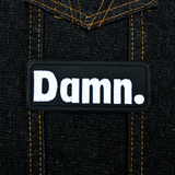 Rectangular black patch with white embroidery spelling the word damn with a period at the end. Shown on a denim jacket.