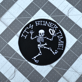 Round black embroidered patch with a dancing skeleton and the words it's boner time in white thread.