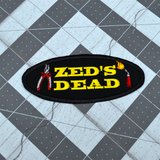 Oval shaped black embroidered patch with the words zed's dead in yellow thread between a pair of pliers and a blowtorch.