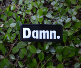 Rectangular black patch with white embroidery spelling the word damn with a period at the end.