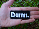 Rectangular black patch with white embroidery spelling the word damn with a period at the end. Shown in the palm of a hand for size reference.