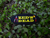 Oval shaped black embroidered patch with the words zed's dead in yellow thread between a pair of pliers and a blowtorch.
