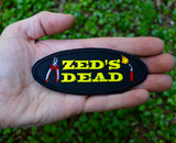 Oval shaped black embroidered patch with the words zed's dead in yellow thread between a pair of pliers and a blowtorch. Shown in the palm of a hand for size reference.