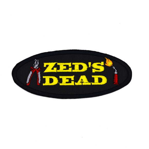 Oval shaped black embroidered patch with the words zed's dead in yellow thread between a pair of pliers and a blowtorch.