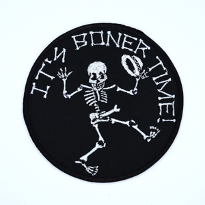 Round black embroidered patch with a dancing skeleton and the words it's boner time in white thread.