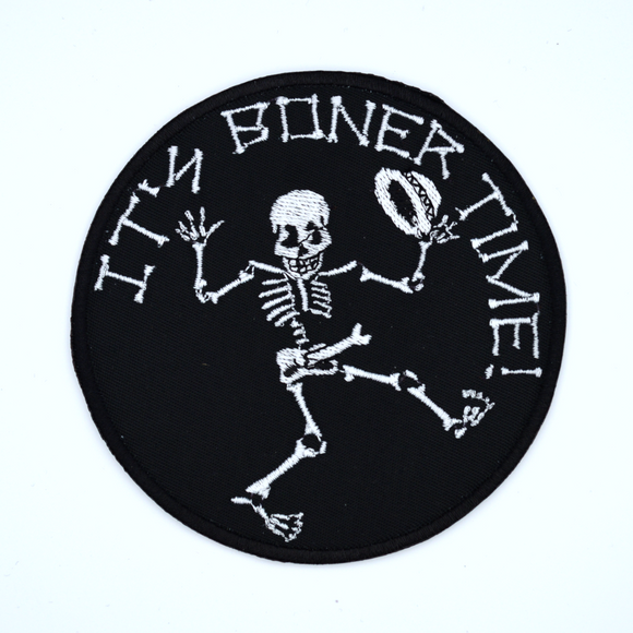 Round black embroidered patch with a dancing skeleton and the words it's boner time in white thread.
