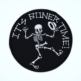 Round black embroidered patch with a dancing skeleton and the words it's boner time in white thread.