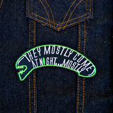 A black and green embroidered patch shaped like an alien head with the words, they mostly come at night, in grey thread. Shown on a denim jacket.