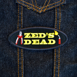 Oval shaped black embroidered patch with the words zed's dead in yellow thread between a pair of pliers and a blowtorch. Shown on a denim jacket.