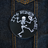 Round black embroidered patch with a dancing skeleton and the words it's boner time in white thread. Shown on a denim jacket.