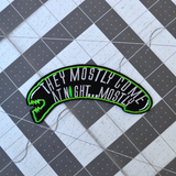 A black and green embroidered patch shaped like an alien head with the words, they mostly come at night, in grey thread.