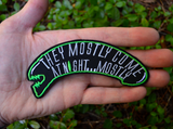 A black and green embroidered patch shaped like an alien head with the words, they mostly come at night, in grey thread. Shown in the palm of a hand.