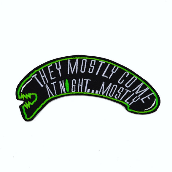 A black and green embroidered patch shaped like an alien head with the words, they mostly come at night, in grey thread.