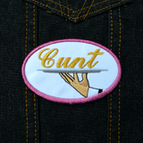 Oval-shaped embroidered patch with a pink border on white fabric showing a hand holding a serving platter with the word cunt on it, sewed in gold thread. Shown on a denim jacket.