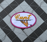 Oval-shaped embroidered patch with a pink border on white fabric showing a hand holding a serving platter with the word cunt on it, sewed in gold thread.