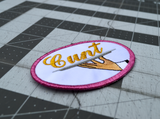 Close up detail of an oval-shaped embroidered patch with a pink border on white fabric showing a hand holding a serving platter with the word cunt on it, sewn in gold thread.