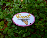 Oval-shaped embroidered patch with a pink border on white fabric showing a hand holding a serving platter with the word cunt on it, sewed in gold thread.