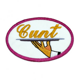 Oval-shaped embroidered patch with a pink border on white fabric showing a hand holding a serving platter with the word cunt on it, sewed in gold thread.
