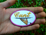 Oval-shaped embroidered patch with a pink border on white fabric showing a hand holding a serving platter with the word cunt on it, sewed in gold thread. Shown in the palm of a hand for size reference.