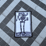 Rectangular black and white embroidered patch of a smoking skeleton in a medieval woodcut design style with the phrase doeth whatever spelled out in old English lettering.