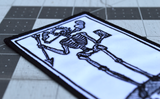 Close up detail of a rectangular black and white embroidered patch of a smoking skeleton in a medieval woodcut design style with the phrase doeth whatever spelled out in old English lettering.