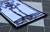 Close up detail of a rectangular black and white embroidered patch of a smoking skeleton in a medieval woodcut design style with the phrase doeth whatever spelled out in old English lettering.
