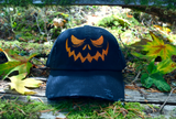 A black baseball style hat with a pumpkin jack o lantern face embroidered on the front in orange thread.