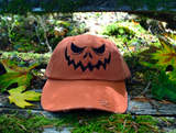 An orange baseball style hat with a pumpkin jack o lantern face embroidered on the front in black thread.