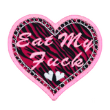 A heart-shaped pink and white embroidered patch on zebra print fabric with the phrase Eat My Fuck.