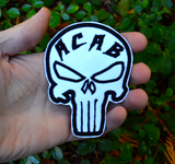 Black and white embroidered Punisher symbol patch with the letters a, c, a, b across the forehead. Held in the palm of a hand.