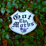 A white badge-shaped embroidered patch with silver border, purple flowers and the phrase, got the morbs, written in old english typesetting.