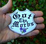 A white badge-shaped embroidered patch with silver border, purple flowers and the phrase, got the morbs, written in old english typesetting. Shown in the palm of a hand for size reference.