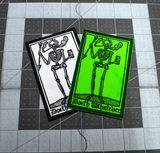 A pair of rectangular embroidered patches in two color variations of a smoking skeleton in a medieval woodcut design style with the phrase doeth whatever spelled out in old English lettering. Shown in a hand for size reference.