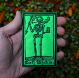 Rectangular lime green and dark green embroidered patch of a smoking skeleton in a medieval woodcut design style with the phrase doeth whatever spelled out in old English lettering. Shown in a hand for size reference.