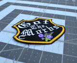 A black badge-shaped embroidered patch with gold border, purple flowers and the phrase, got the morbs, written in old english typesetting.