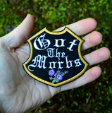 A black badge-shaped embroidered patch with gold border, purple flowers and the phrase, got the morbs, written in old english typesetting. Shown in the palm of a hand for size reference.