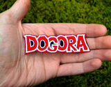 A red, black, and white embroidered logo patch for Dogora, the classic Kaiju monster movie. Shown in the palm of a hand for size reference.