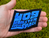 Blue and black embroidered logo patch for the classic kaiju monster movie, Yog, Monster from Space. Shown in the palm of a hand for size reference.