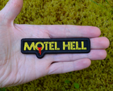 Rectangular black and yellow embroidered logo patch for the classic comedy horror slasher movie Motel Hell. Shown in the palm of a hand for size reference.