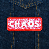 A rectangular red bordered embroidered patch on white fabric with the word chaos carved out of a web of random lines of red thread. Shown on a denim vest.