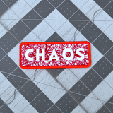 A rectangular red bordered embroidered patch on white fabric with the word chaos carved out of a web of random lines of red thread.