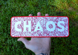 Rectangular embroidered patch with the word chaos cut out of random lines of red thread on white fabric. Shown in the palm of a hand for size reference.