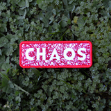 A rectangular red bordered embroidered patch on white fabric with the word chaos carved out of a web of random lines of red thread.