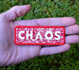 A rectangular red bordered embroidered patch on white fabric with the word chaos carved out of a web of random lines of red thread. Shown in the palm of a hand for size reference.