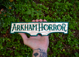 Rectangular embroidered patch resembling a wooden sign with the logo of the Arkham Horror board and card games. Shown being held in a hand for size reference.