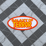 An oval-shaped white, orange, and yellow embroidered patch made from the title logo of the cult classic sci-fi horror movie Galaxy of Terror.