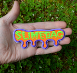 A purple, orange, and neon green embroidered patch shaped like dripping ooze with the word slimebag. Shown in the palm of a hand for size reference.