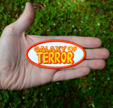 An oval-shaped white, orange, and yellow embroidered patch made from the title logo of the cult classic sci-fi horror movie Galaxy of Terror. Shown in the palm of a hand for size reference.