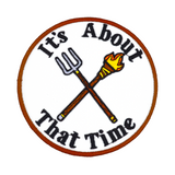 Circular embroidered patch with a brown border, a torch and pitchfork crossed over each other in the middle, surrounded by the phrase, it's about that time.