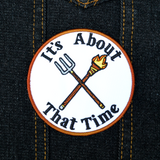 Circular embroidered patch with a brown border, a torch and pitchfork crossed over each other in the middle, surrounded by the phrase, it's about that time.